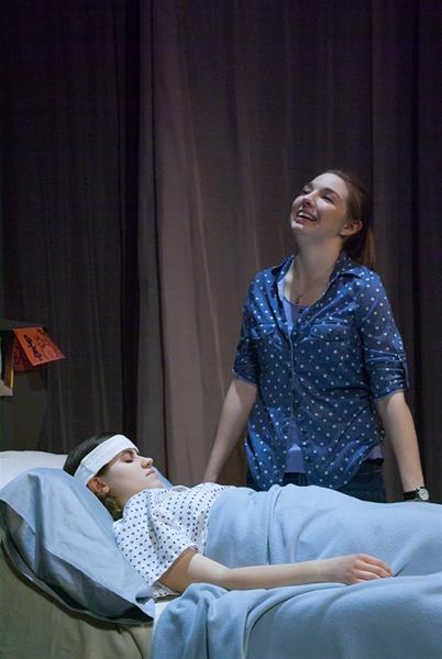 A person lies in a hospital bed with a bandage on their head, covered with a blue blanket. Another person stands beside the bed, smiling and looking upward. They are wearing a blue polka dot shirt. The scene is dimly lit.