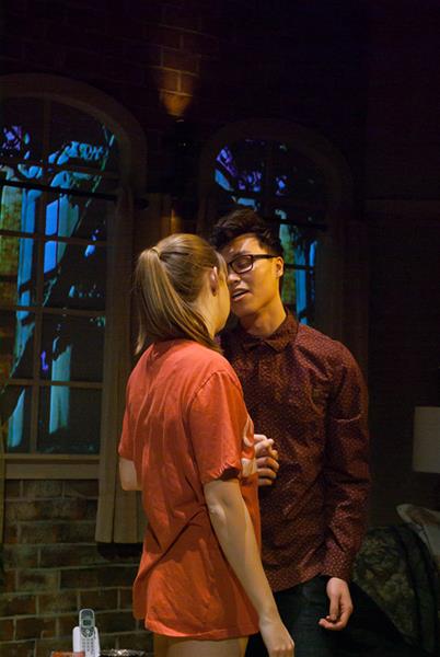 A man and woman stand facing each other in a dimly lit room with brick walls and arched windows. The woman has her back to the camera, while the man faces her, wearing glasses and a patterned shirt. A remote control is on a nearby table.