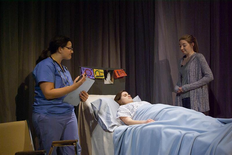 A healthcare worker in scrubs holds a clipboard and stands beside a patient lying in a hospital bed. Another person stands next to the bed, appearing concerned. The setting suggests a hospital room.