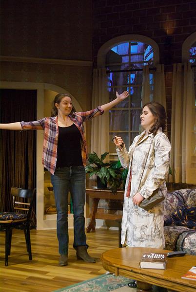 Two women are in a room; one wearing a plaid shirt and jeans gestures with arms wide open, while the other in a patterned robe holds an object. They stand near a small table with a book, chair, and a plant in the background.