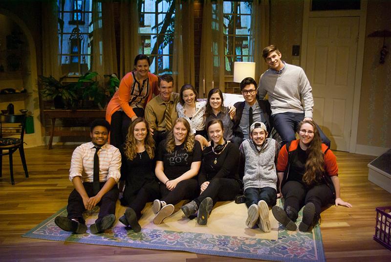 A group of eleven people sits and kneels on a stage, smiling for the camera. They are casually dressed and appear to be in a warmly lit room with plants and large windows in the background.