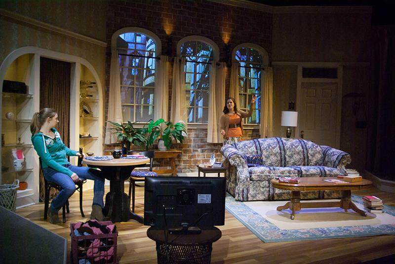 A living room set on stage with two women. One woman sits at a round table, wearing a teal sweater, and the other stands near a window, gesturing with one hand. The room is furnished with a floral couch, coffee table, and decorated with plants.