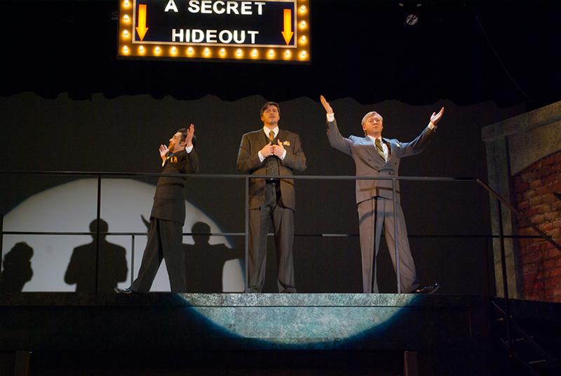 Three men in suits stand on a balcony stage under a sign reading "A Secret Hideout." They are striking theatrical poses with their arms raised. A spotlight casts their shadows on the wall behind them.