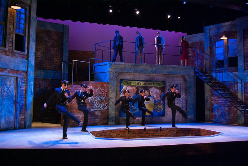 Actors in police costumes perform a choreographed dance on stage with a cityscape backdrop. The set includes graffiti-covered walls, stairs leading to an upper level, and dramatic lighting that enhances the urban atmosphere.