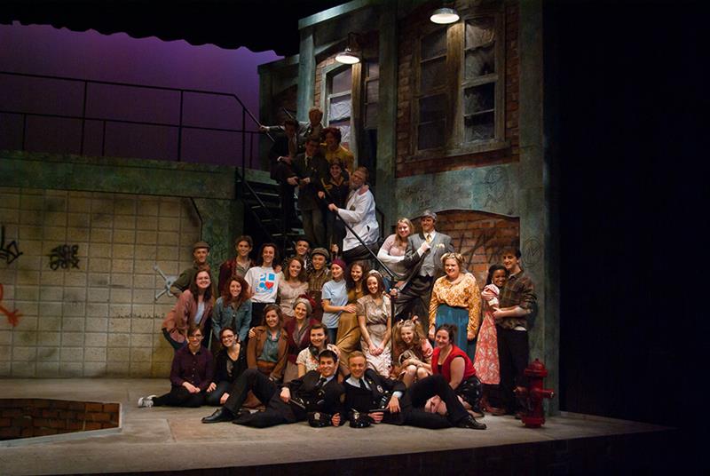 A diverse group of people pose on a stage set resembling an urban alleyway, with graffiti and brick walls. Some sit on the stairs, others stand or sit on the ground, smiling. The lighting is warm, giving a theatrical atmosphere.