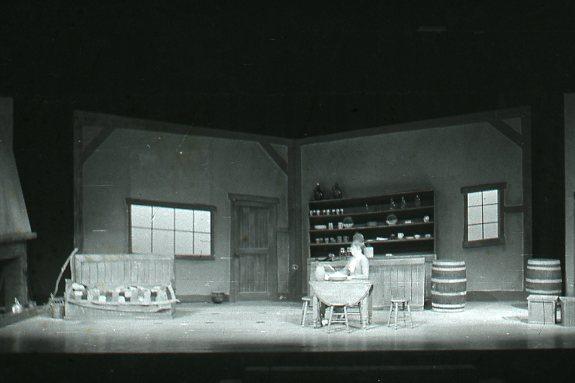 A black and white photo of a stage set depicting a rustic room. It includes a window, a wooden door, shelves with pottery, barrels, a table with a chair, and a bench. The lighting casts shadows, creating a moody atmosphere.