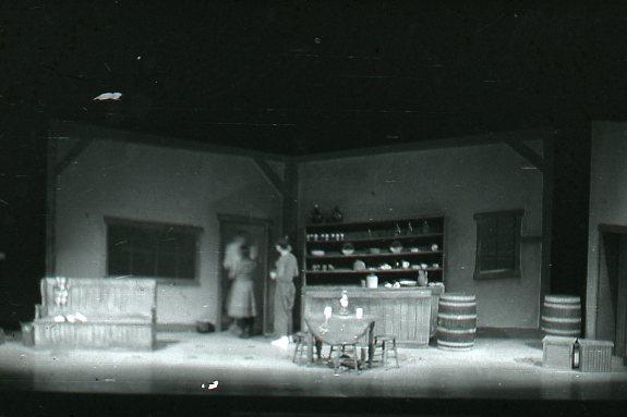 A black-and-white image of a stage set depicting a rustic interior. It features wooden furniture, a table with chairs, shelves stocked with dishes, and barrels. Two blurred figures are near a door, contributing to the scene's atmospheric depth.