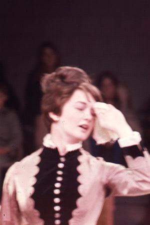 A person with short hair, wearing a vintage-style outfit with buttons down the front, wipes their forehead with a cloth. The background is blurred, showing indistinct figures. The person appears to be in motion or expressing emotion.