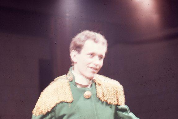 A person in a green costume with gold fringe on the shoulders, stands under stage lights. The background is dark and out of focus.