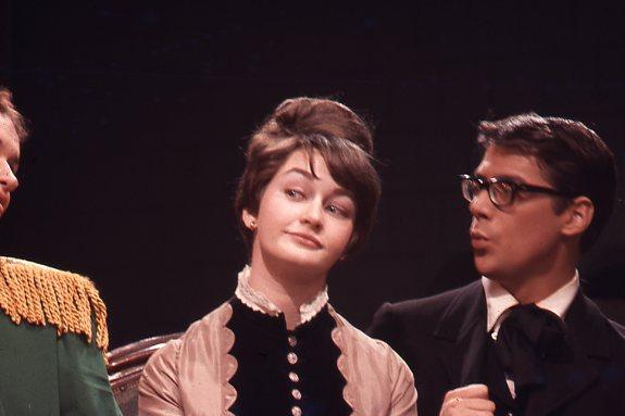 A woman with an updo hairstyle and a vintage dress looks to her right with a playful expression. A man with glasses, wearing a dark suit and bow tie, appears to be speaking to her. The background is dark, creating a theatrical atmosphere.