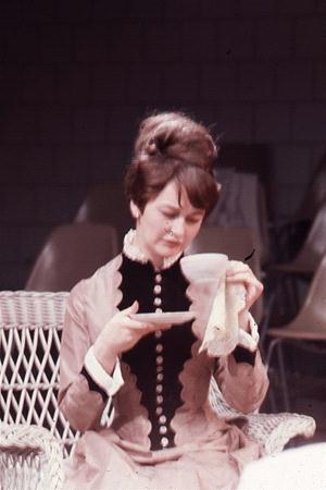 A woman in a vintage dress with decorative buttons sits on a wicker chair. She is holding a teacup and saucer, gazing at the cup. Her hair is styled in an updo. The background features a few empty chairs.