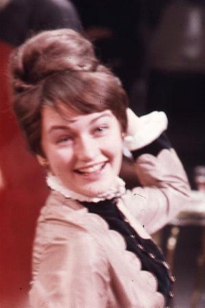 A woman with brown hair styled in an updo smiles and looks over her shoulder. She wears a vintage-inspired outfit with a lace collar and buttons, set against a blurred background.