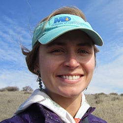 A person wearing a green visor and a textured jacket smiles at the camera. The background shows a landscape with sparse vegetation and a partly cloudy sky.