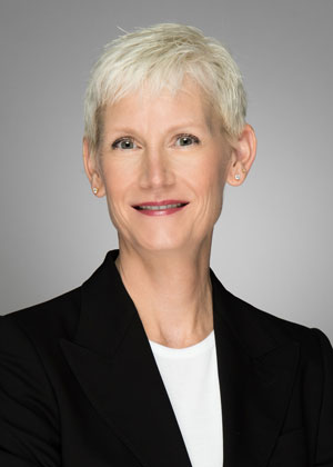 A person with short, white hair wearing a black blazer over a white top is smiling at the camera. The background is a plain, neutral grey.