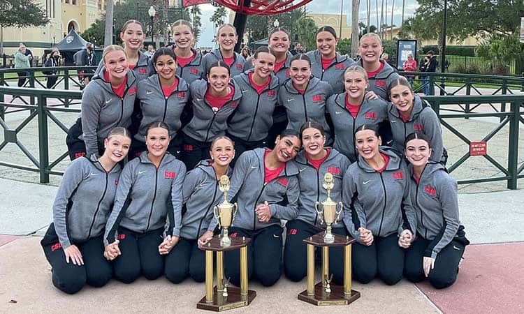 A group of 18 people dressed in matching gray athletic jackets pose outdoors with two trophies on the ground in front of them. They are smiling and kneeling or standing in rows, with a decorative structure and greenery visible in the background.