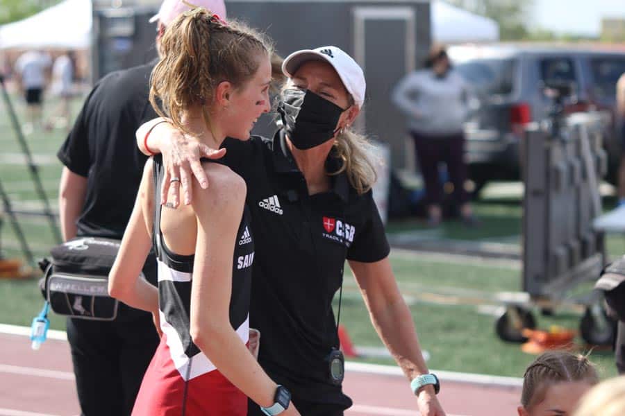 A coach, wearing a black mask and black athletic gear, gives an encouraging pat to a young female athlete dressed in a red and black track uniform, standing on a track field. The background includes other athletes, equipment, and a parked vehicle.