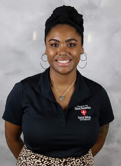 A person is standing in front of a grey background smiling. They are wearing a black shirt with the text "College of Saint Benedict Saint John’s University" and a headscarf. They have hoop earrings, a nose ring, and a necklace.