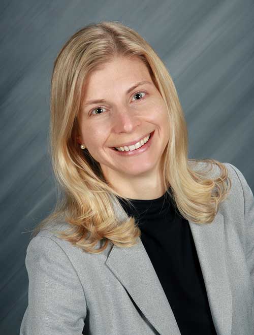 A woman with long blonde hair smiles at the camera. She is wearing a light gray blazer over a black top. The background is a grayish gradient.