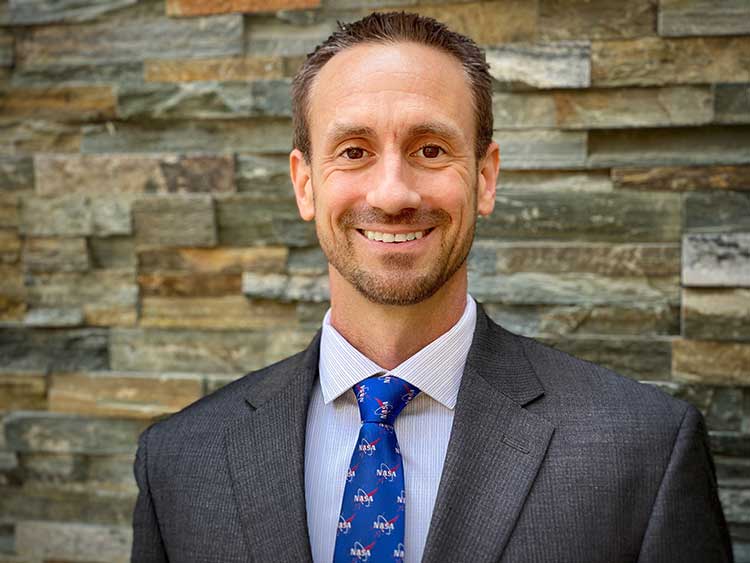 A man in a dark suit, light blue shirt, and NASA-themed tie smiles at the camera. He stands in front of a textured stone wall.