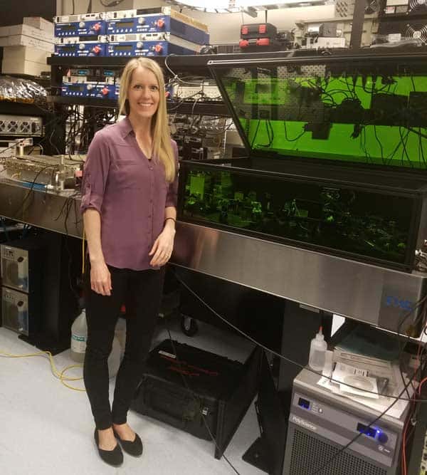 A woman with long blonde hair, wearing a purple blouse and black pants, stands next to a large lab machine with wires and components. She is in a research lab setting, filled with equipment and technology.