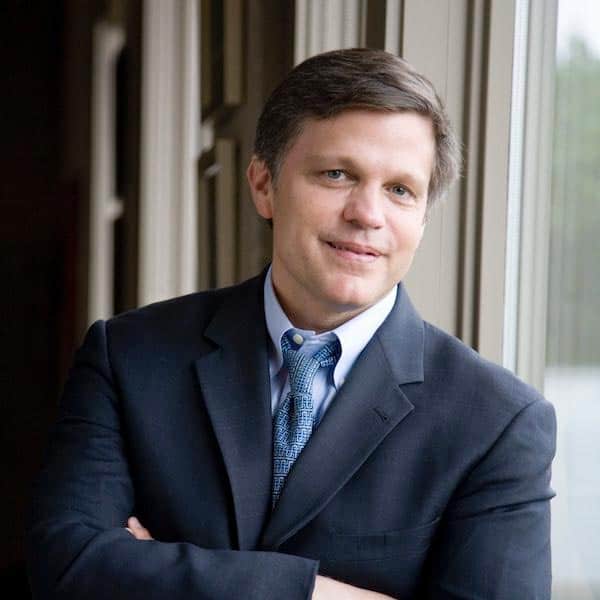 A person in a dark suit, light blue shirt, and patterned blue tie stands with arms crossed, leaning against a tall window. The person has short brown hair and is smiling softly. The background is softly lit, and greenery is visible outside the window.