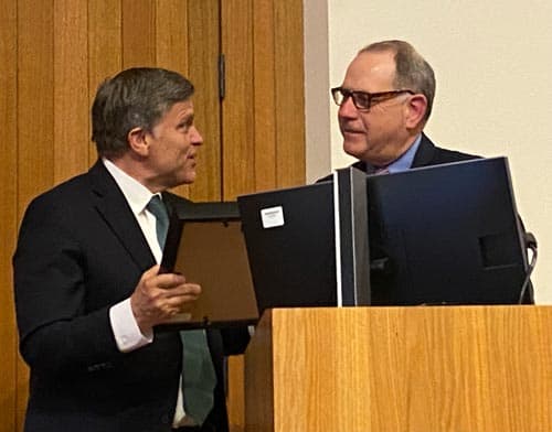 Two men in suits are standing behind a wooden podium. One man, holding a framed certificate, is speaking to the other man, who is wearing glasses and standing next to a computer monitor. They appear to be in a formal setting, such as a conference or award ceremony.