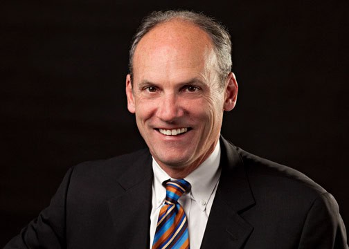 A smiling bald man wearing a black suit, white shirt, and multicolored striped tie, is pictured against a dark background. He is looking directly at the camera.