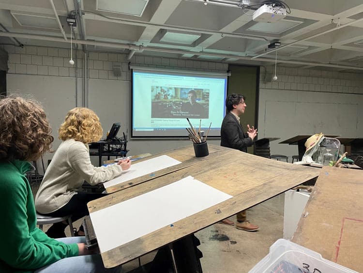 A person standing and speaking in front of a classroom with a projector screen behind them, displaying a presentation. Two students, seated at desks with drawing materials, listen attentively. The room has art supplies and is brightly lit.