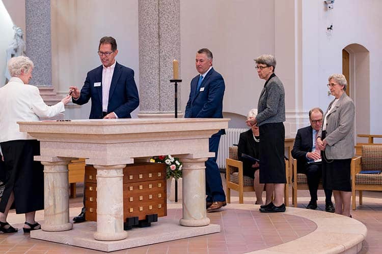 A group of people, including men in suits and women in formal attire, stand around a stone altar in a church. Some are seated. The setting appears solemn, possibly for a religious or memorial service.