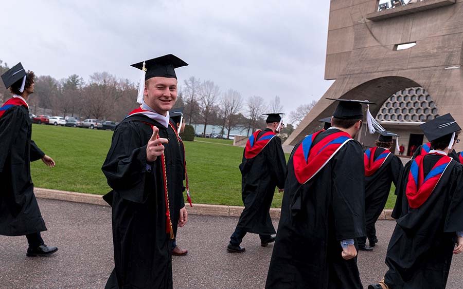 A group of graduates in black caps and gowns with red and blue stoles walk outdoors on a cloudy day. One of them is smiling and pointing at the camera. An architectural structure with circular designs is visible in the background.