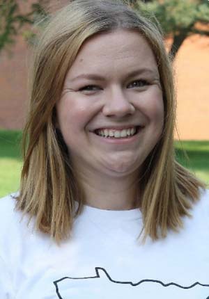 A smiling person with shoulder-length blonde hair is standing outdoors. They are wearing a white shirt with a simple outline design. The background includes greenery and what appears to be a brick building.