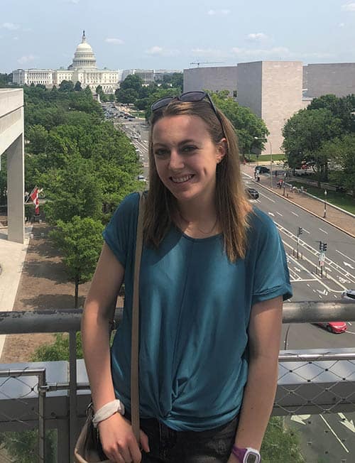 A woman with shoulder-length brown hair stands smiling on a balcony overlooking a city street. She is wearing a teal blouse, sunglasses on her head, and a light brown crossbody bag. In the background is a dome-shaped building surrounded by trees and other structures.