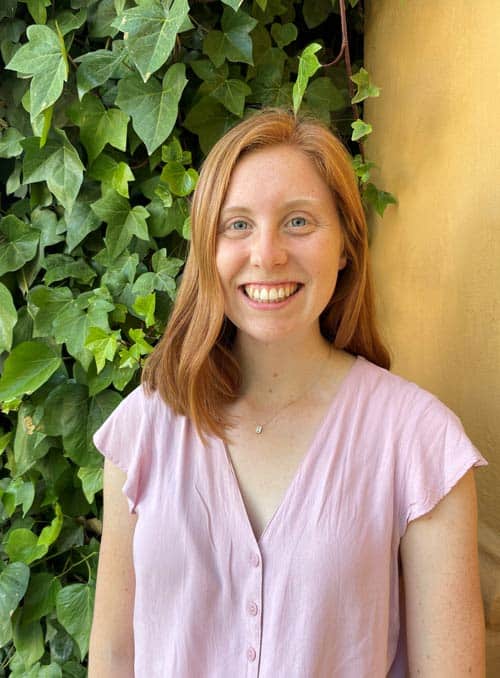 A smiling person with shoulder-length red hair stands in front of a wall covered with green ivy leaves. They are wearing a light pink, short-sleeved blouse.