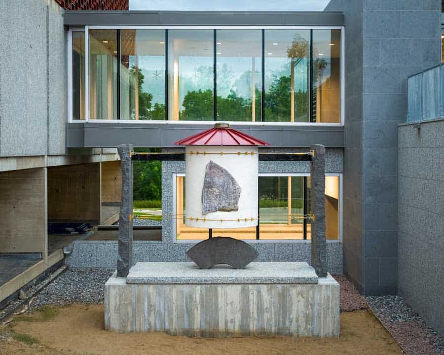 A modern building features a large glass window facade on the second floor. Below, a central art installation with stone pillars supports a suspended artwork, and a stone bench is in front. The scene is set against a backdrop of greenery.