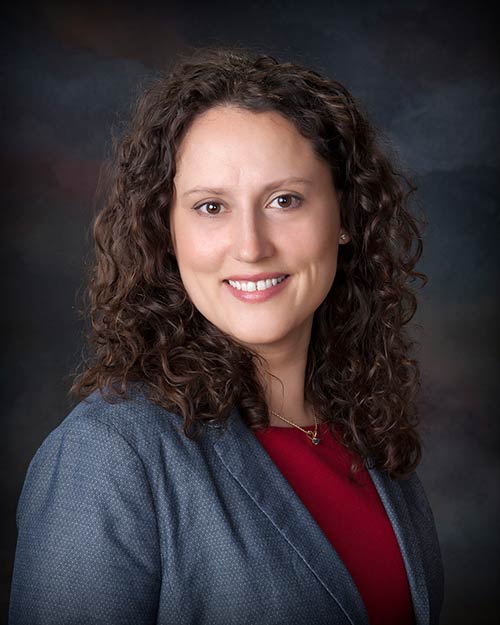 A woman with curly brown hair wearing a grey blazer over a red top smiles at the camera. The background is a dark, blurred gradient.