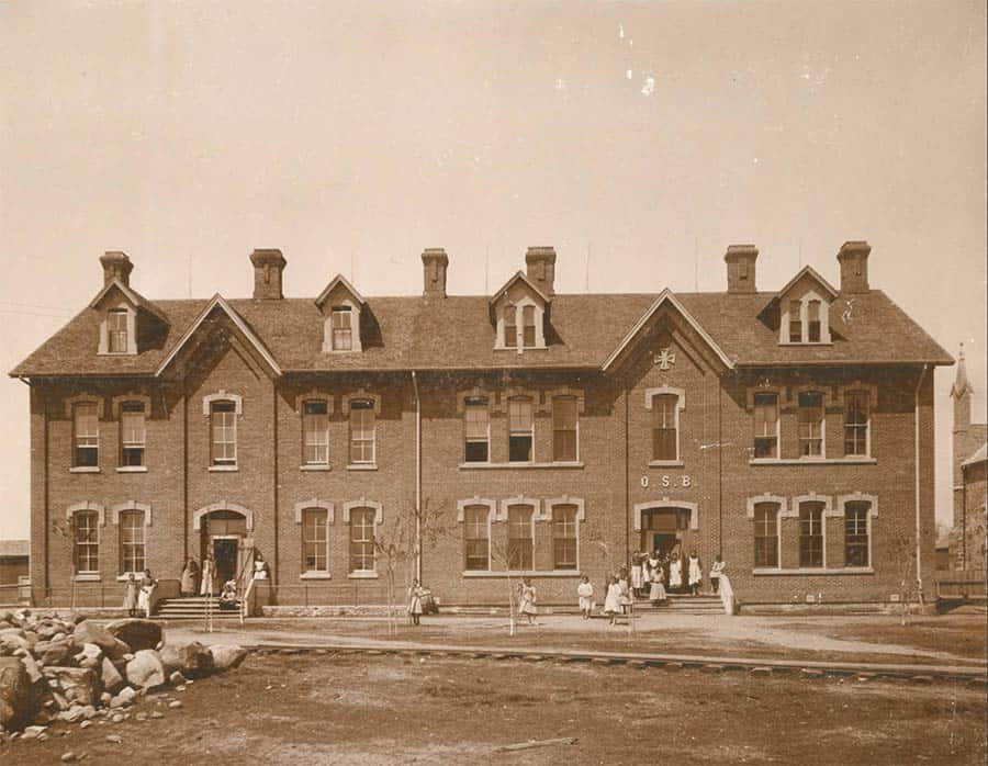 A sepia-toned photograph of a large, two-story brick building with numerous windows and gabled roofs. Several people are visible in front of the building, some standing and some seated. The building bears the letters "O.S.E." above the entrance.