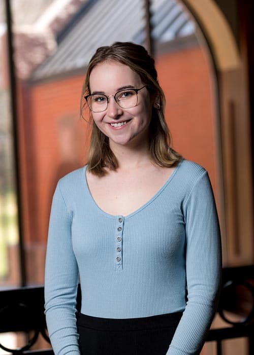 A woman with glasses and shoulder-length hair smiles at the camera. She is wearing a long-sleeved, ribbed light blue top with buttons and black pants. The background shows a red brick building and arched windows, slightly blurred.