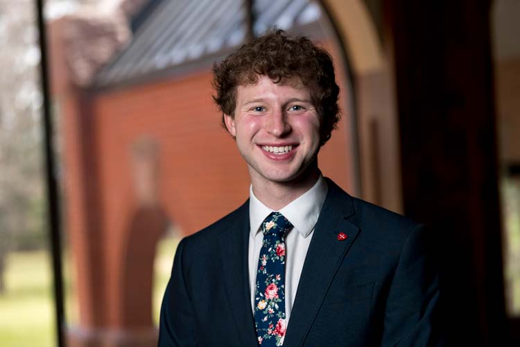 A young man with curly hair and a bright smile stands in front of a blurred background featuring a red brick building. He is wearing a dark suit, white dress shirt, and a floral tie.