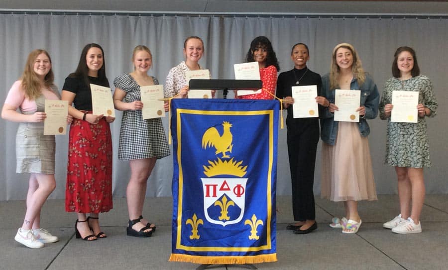 Eight individuals stand on a stage holding certificates, smiling at the camera. They are positioned behind a blue and yellow banner featuring a bird emblem and the Greek letters ΠΔΦ. The backdrop is a plain gray curtain.