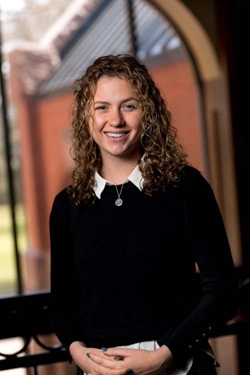 A person with curly hair smiles at the camera while standing indoors. They are wearing a black sweater over a white collared shirt and a round pendant necklace. A window with a view of brick architecture is visible in the background.