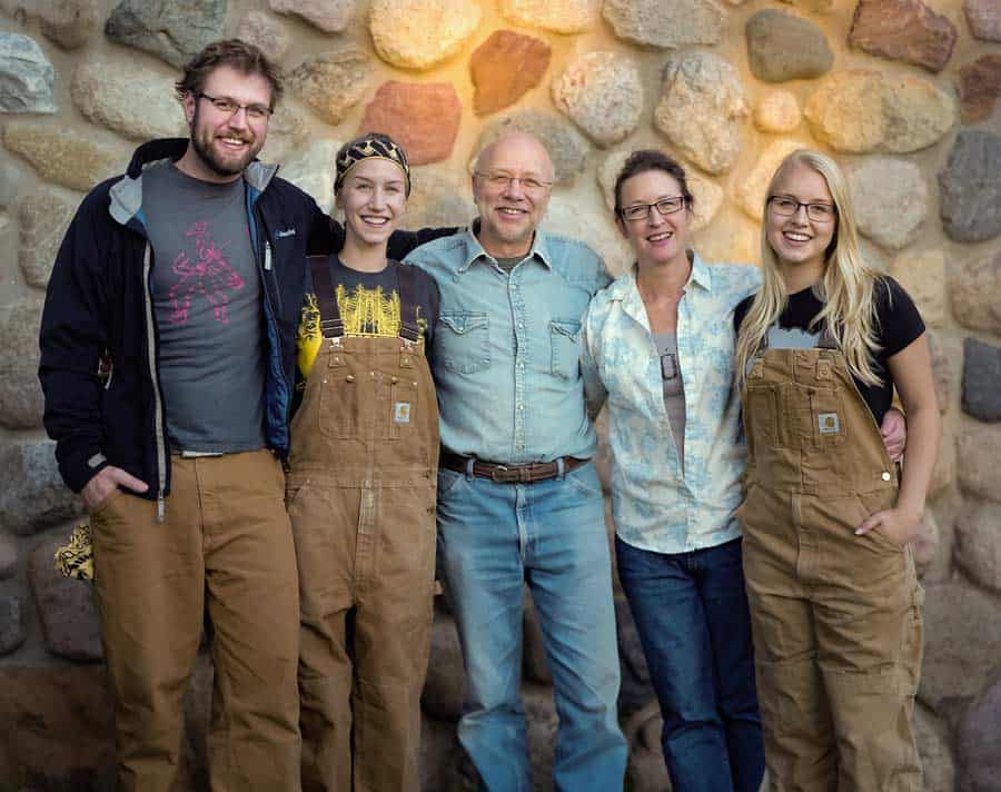 A group of five people, including two men and three women, stand side by side smiling in front of a stone wall. Two of the women and one man are wearing matching brown overalls, and the other two individuals are dressed in casual clothing.