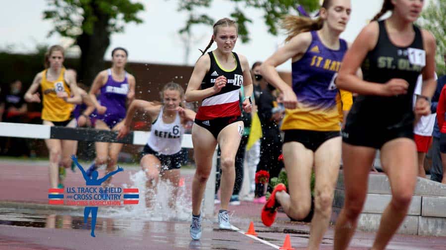 A group of female athletes competes in a track and field race, with one athlete running through a water obstacle. The U.S. Track & Field and Cross Country Coaches Association logo is visible in the bottom left corner. The athletes wear various team uniforms.