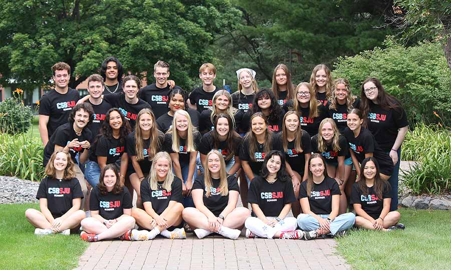 A group of 29 young adults, wearing matching black t-shirts with "CSB/SJU" printed on them, pose together outdoors. They are sitting and standing on a pathway surrounded by greenery, smiling at the camera on a slightly overcast day.