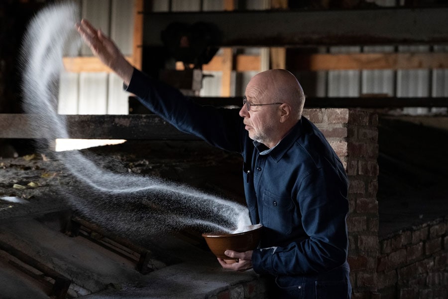 A man with a bald head and glasses, wearing a dark blue shirt, stands inside a rustic building and throws a handful of white powder into the air from a wooden bowl. The powder forms an arc as it disperses.