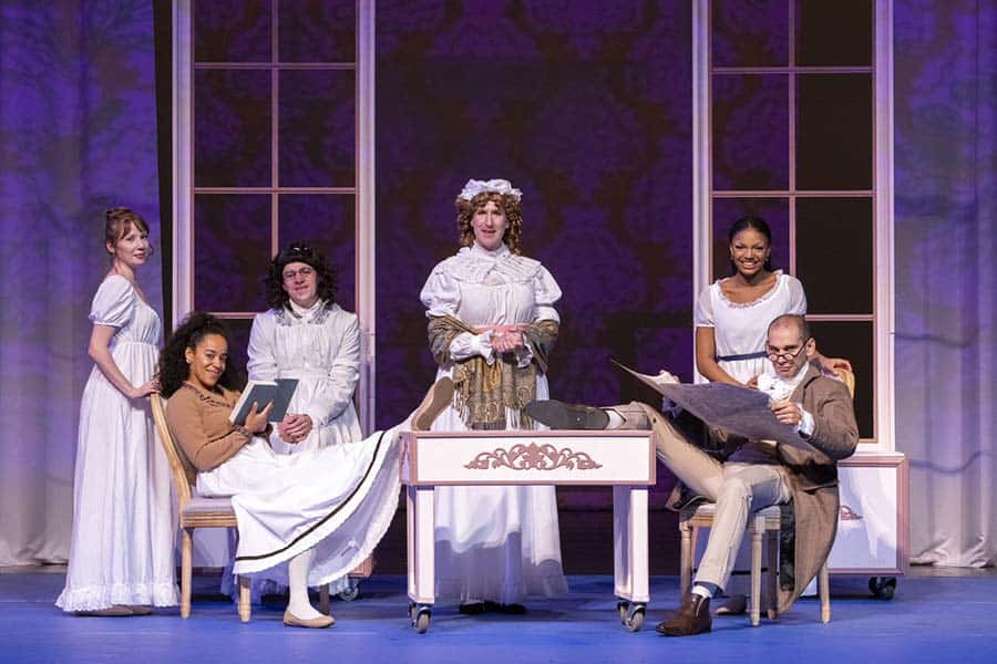 Six actors are on stage dressed in Regency-era costumes. Three women stand behind, while three others sit in front. One man reads a newspaper; another holds a book. The set features two large window frames, evoking an early 19th-century interior.