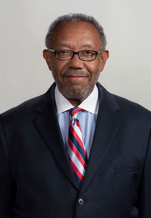 A man with short gray hair and a gray beard, wearing glasses, a dark suit jacket over a light blue shirt, and a red, white, and blue striped tie stands against a plain background. He is looking directly at the camera with a neutral expression.