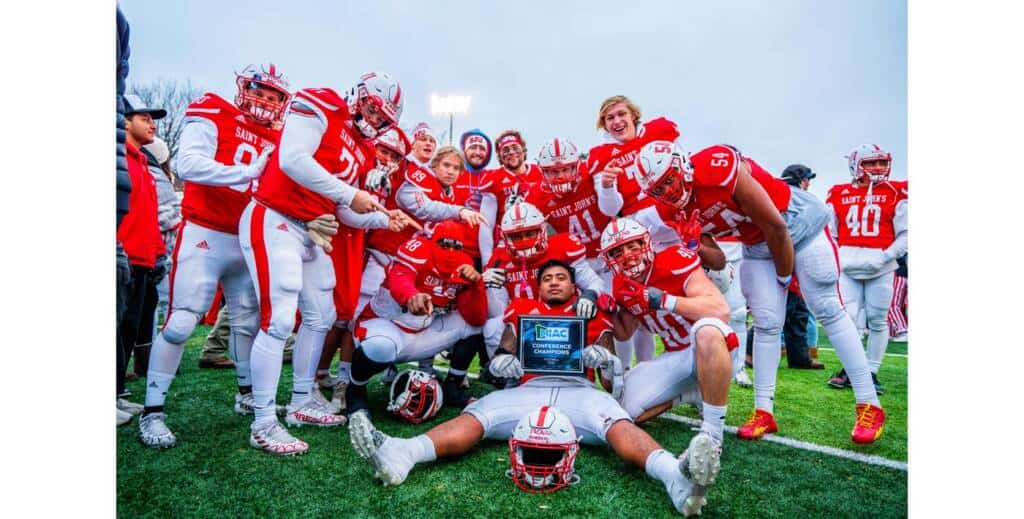 A football team in red and white uniforms celebrates on the field. Players are gathered around, some standing and others kneeling. A player lying on the ground holds a championship plaque, while teammates cheer and point at him. They are smiling joyfully.