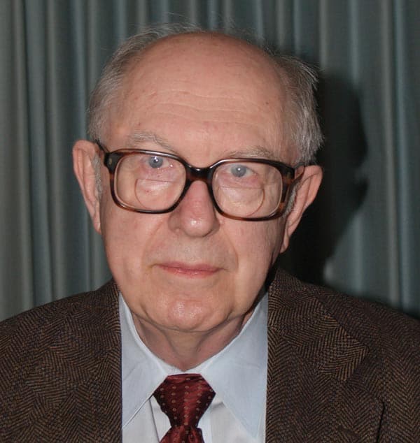 An elderly man with glasses and thinning hair wears a brown suit, white shirt, and red tie. He is standing in front of a curtain with a neutral expression.