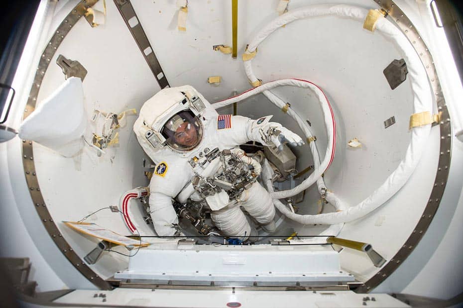 An astronaut in a white spacesuit with an American flag on the sleeve is inside a module, surrounded by various cables and equipment. The astronaut appears to be floating or in a state of weightlessness. The background is mostly white with metallic and fabric elements.