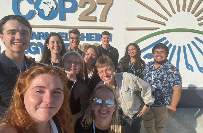 A group of people is smiling and taking a selfie in front of a COP27 sign. The sign features a partial globe graphic and a stylized sun with rays. The group appears to be enjoying themselves on a sunny day.